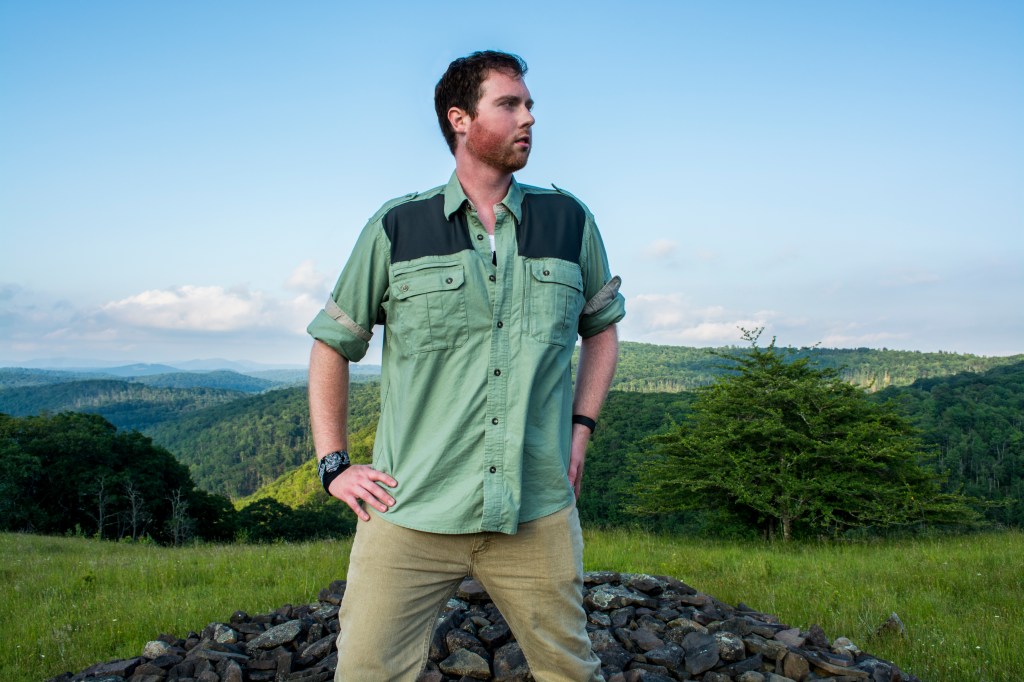 A man in a green shirt standing at the top of a mountain with mountain scenery, trees, grass, and a rock pile in the background.